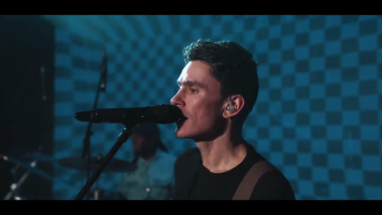 Side view of a man singing into a microphone with a drummer and a checkered blue wall in the background.