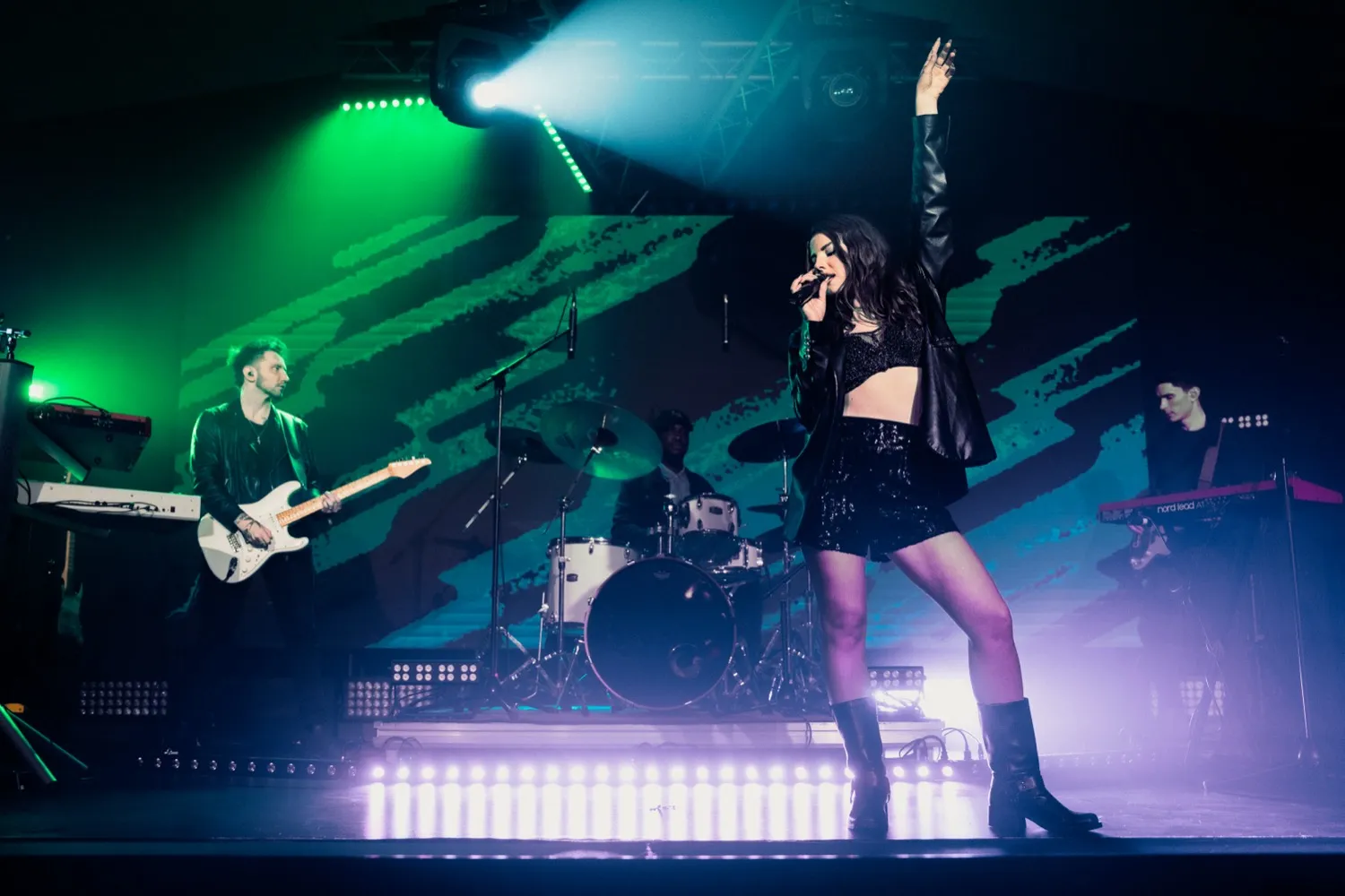Female singer in black outfit performing with band on stage under colored lighting.