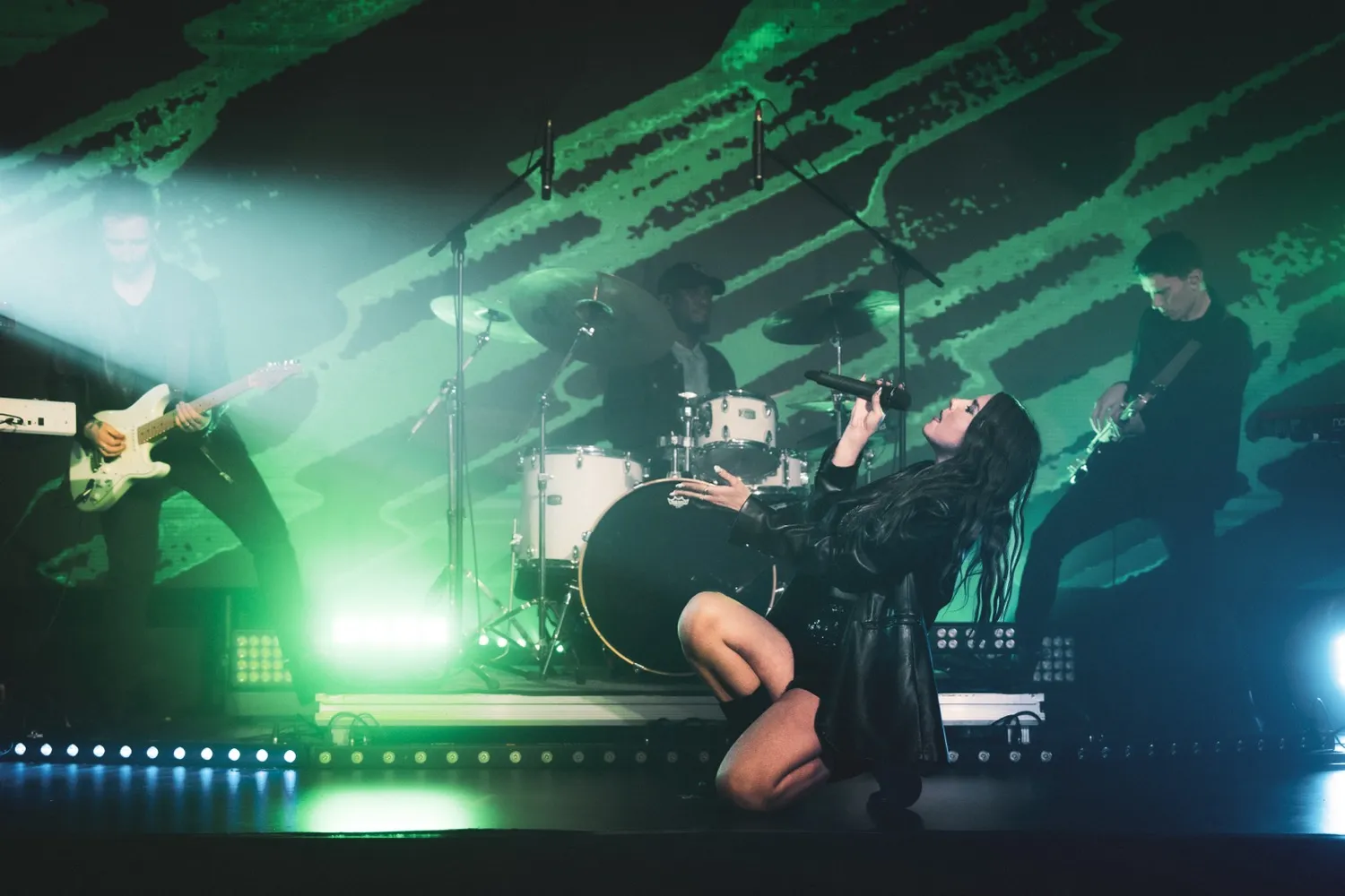 Female singer kneeling on stage passionately performing with band members playing guitar and drums in the background under green lighting.