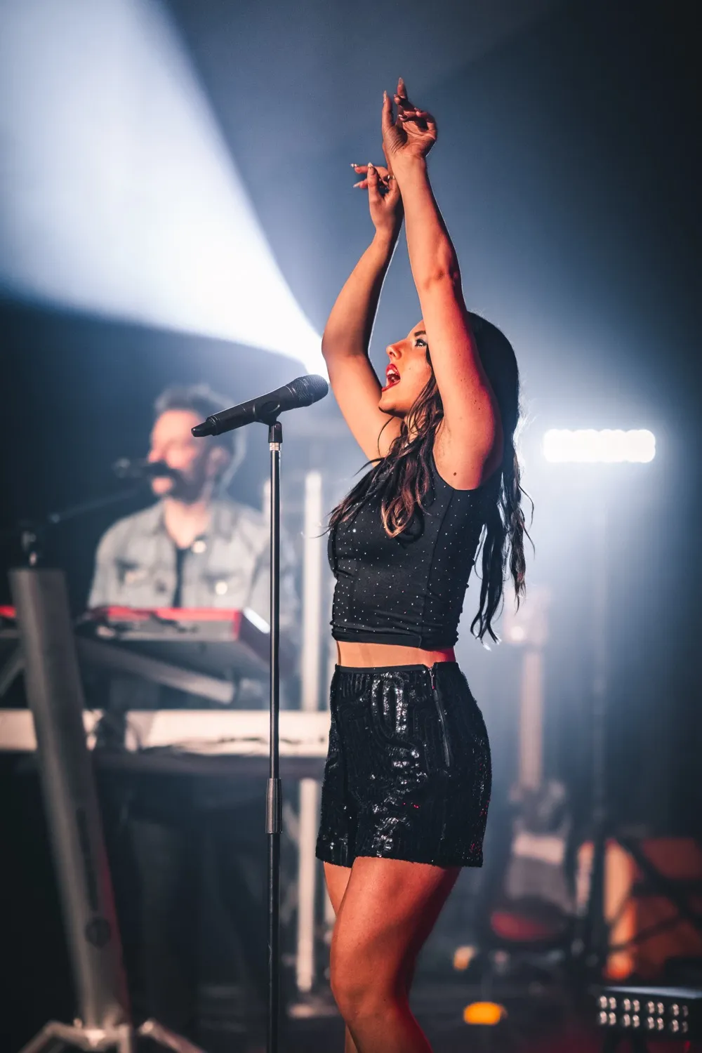 Female singer in black outfit performing passionately on stage with arms raised, illuminated by stage lights.