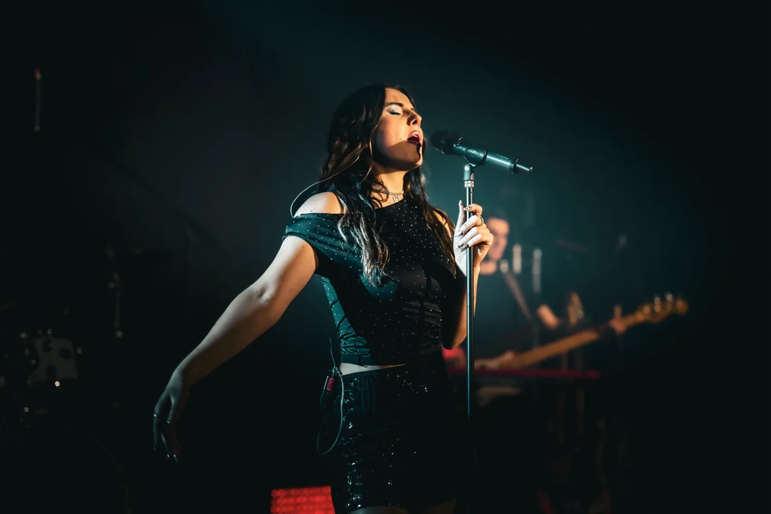 Female singer passionately performing on stage with microphone, wearing a black outfit under spotlight.