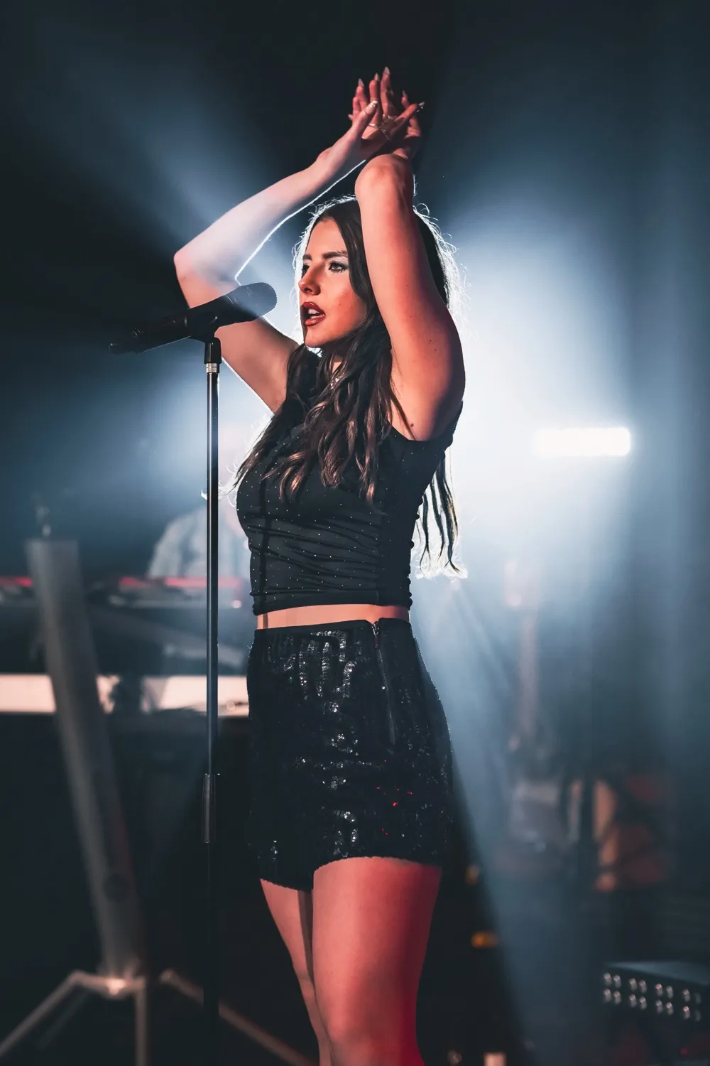 Female singer in black outfit with hands raised, performing on stage with microphone and spotlight.