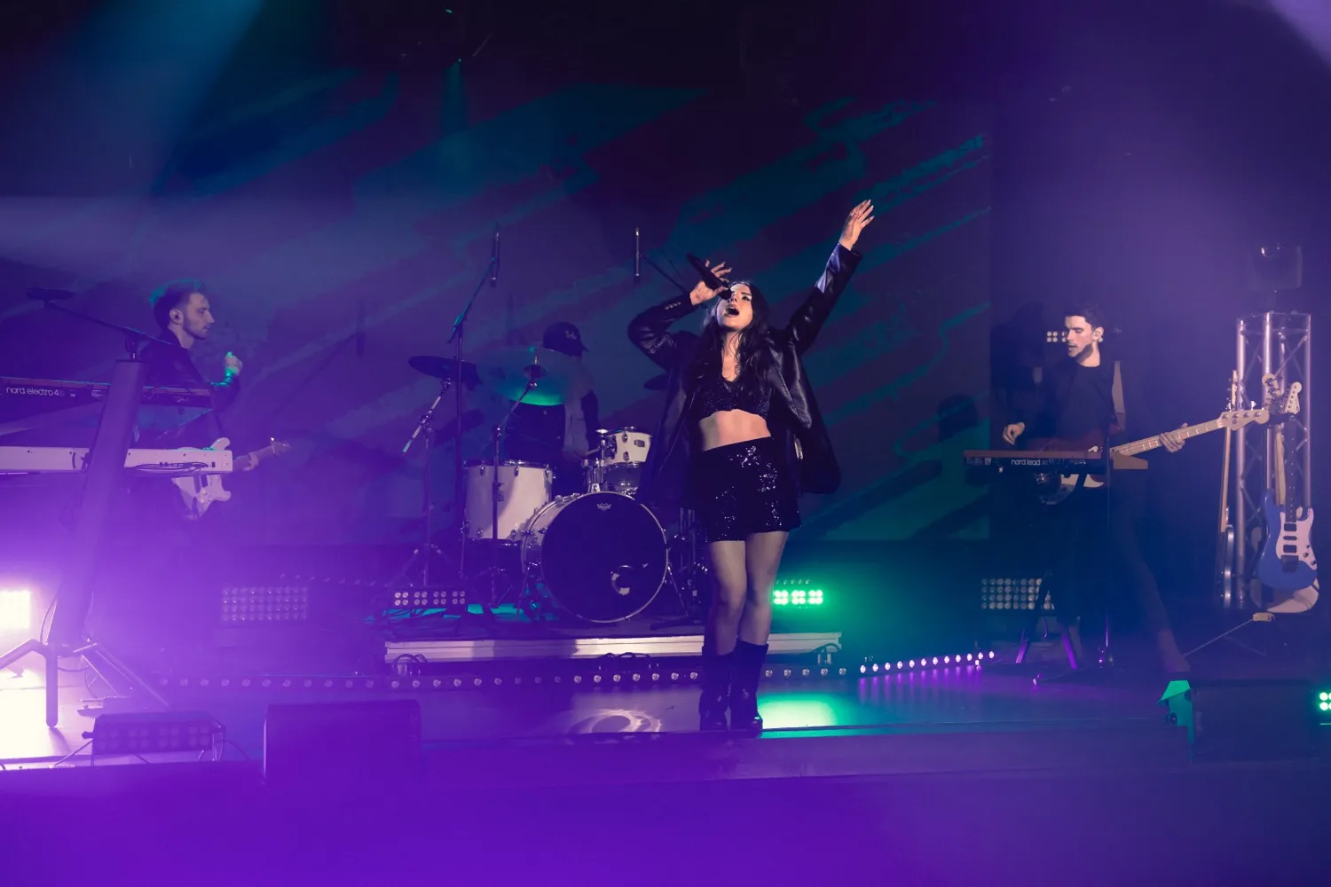 Female singer performing on stage with a band, purple and green stage lighting.